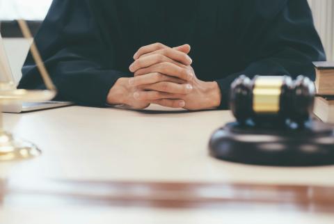 Judge sitting at a desk with their fingers interlocked. A scale and gavel appear on the desk. 